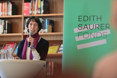 Eine Frau steht in einer Bibliothek vor einem Edith Saurer Fonds Rollup und spricht in ein Mikrofon. © ESF / Christopher Glanzl
