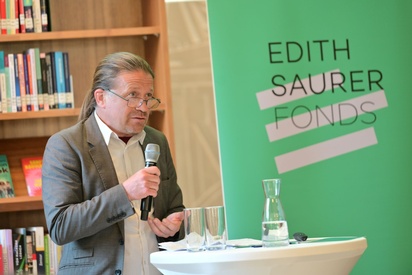 Ein Mann in einem grauen Anzug und einem Mikrofon hält in einer Bibliothek eine Rede. © ESF / Christopher Glanzl