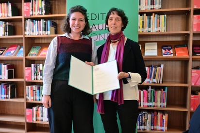 Zwei Frauen stehen vor einem Bücherregal, eine präsentiert eine Urkundenmappen. © ESF / Christopher Glanzl